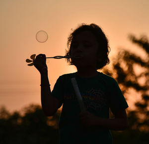 Silhouette girl blowing bubbles against sky during sunset