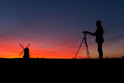 Silhouette photographer with tripod by windmill against sky at dusk