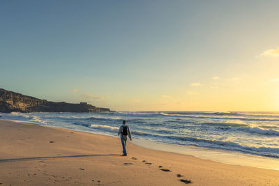 Man walking at beach against sky during sunset