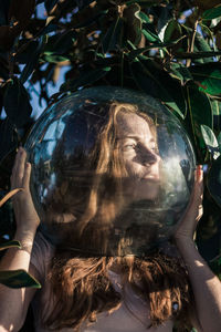 Close-up of young woman wearing glass ball on head against leaves