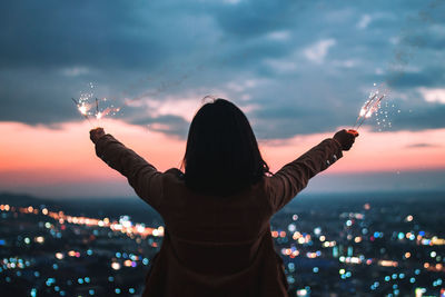 Rear view of woman holding sparklers against illuminated cityscape at dusk