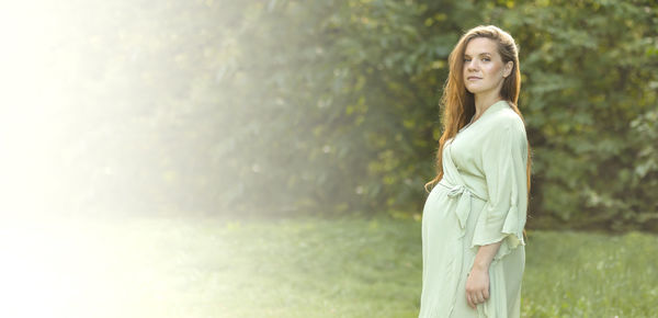 Banner white pretty pregnant woman stands, looks at camera. green trees, park is on background. 