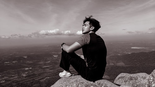 Side view of young man sitting on rock looking at landscape
