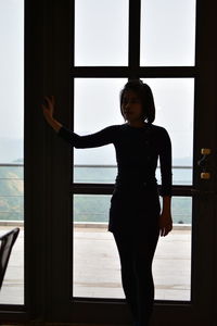 Portrait of young woman standing against window in room