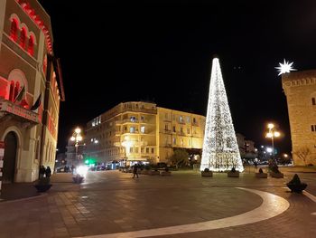 Illuminated christmas tree by buildings against sky at night