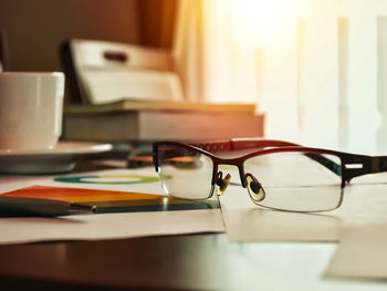 Close-up of eyeglasses with books and cup on table against window