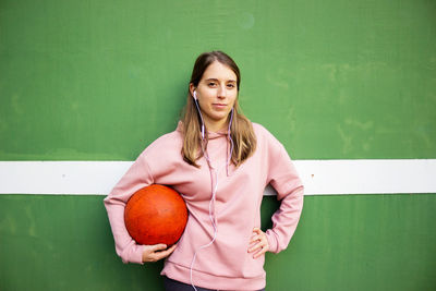 Portrait of young woman holding basketball against wall