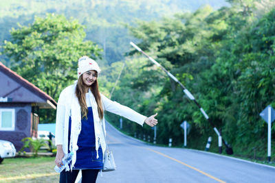 Portrait of smiling young woman hitchhiking on road against trees