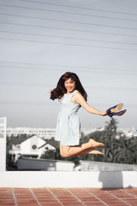 Portrait of cheerful woman jumping against sky at building terrace during sunny day