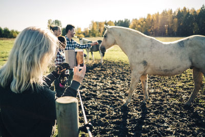 Mature female farmer photographing friends and horse at animal pen