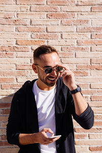 Smiling mid adult man wearing sunglasses using smart phone while standing against brick wall