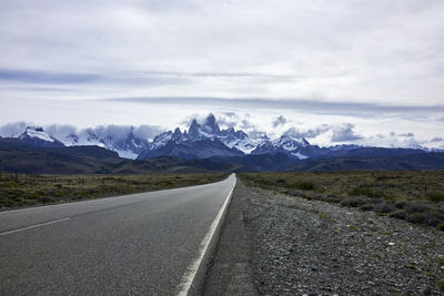 Empty country road leading towards mountains against cloudy sky during winter