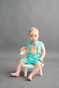 A funny little boy in a summer suit is sitting on a white children's chair on a gray background 