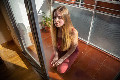 Thoughtful young woman with blond hair kneeling in balcony