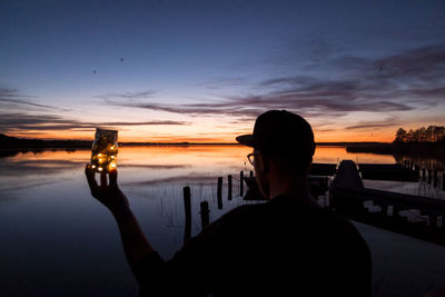 Rear view of man holding illuminated decoration by lake against sky during sunset