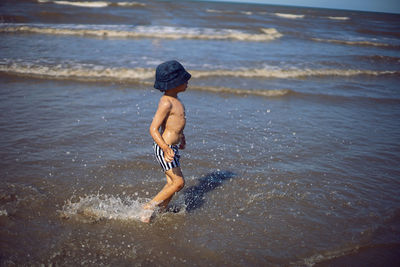 Boy child runs along the beach sea in summer in a panama hat and striped shorts