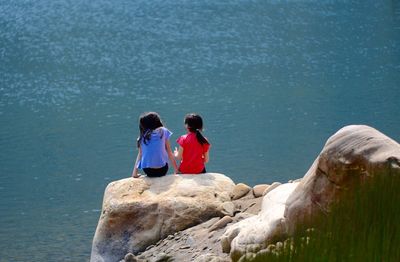 Rear view of girls sitting on rock formation by lake