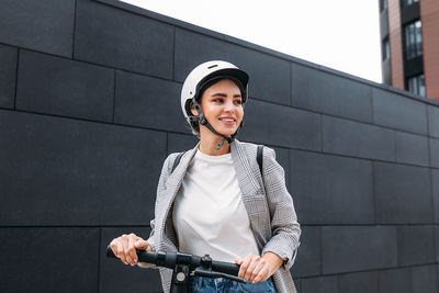 Smiling woman in helmet with an electric scooter at black wall looking away	