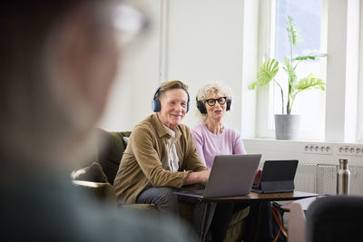 Senior man and woman sitting in living room and using laptop and digital tablet to edit podcast