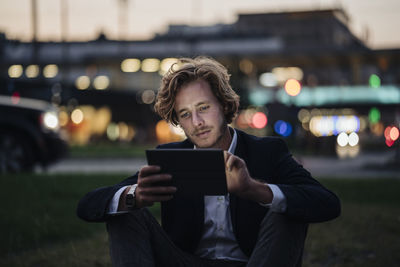 Businessman sitting on meadow at dusk using tablet