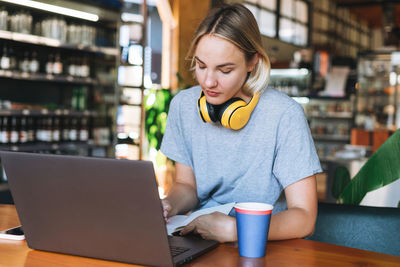 Young smiling blonde woman freelancer with yellow headphones working on notebook on table at cafe