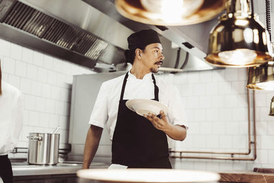 Male chef looking away while holding plate in commercial kitchen