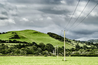 Electric poles on field by mountain against cloudy sky