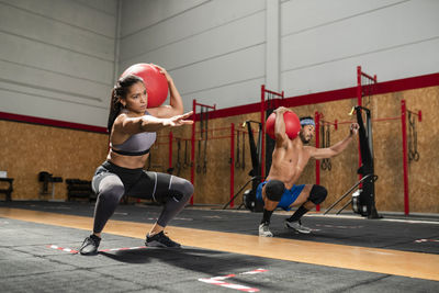 Concentrated sportswoman and sportsman doing squats with heavy medical balls during functional intense training in sports club