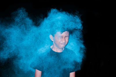 Boy with eyes closed amidst blue powder paint against black background