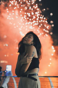 Young woman posing while standing against firework display