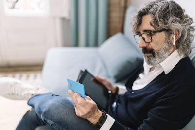 Handsome man looking at credit card holding digital tablet while sitting at home