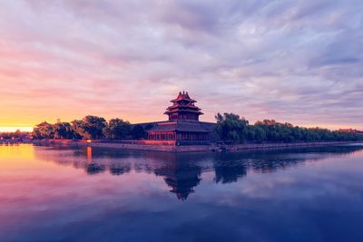 Building by lake against dramatic sky at sunset