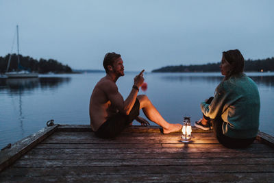 Full length of couple talking while sitting on pier by sea against sky