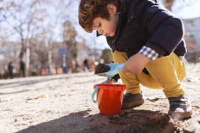 Full length of boy playing with sand pail and shovel while crouching at beach