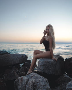 Young woman sitting on rock at sea shore against clear sky during sunset