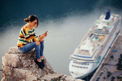 Full length of young woman using phone while sitting on retaining wall against lake