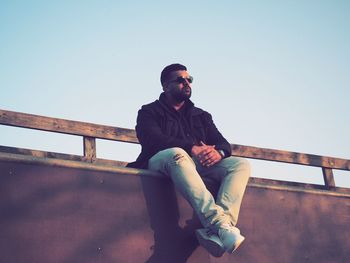 Young man looking away while sitting on railing against clear sky