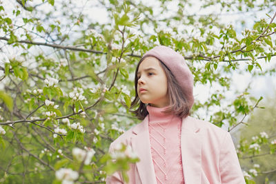 Portrait of little girl stands in a blooming white apple tree in the park.