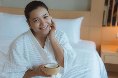 Portrait of smiling beautiful young woman applying moisturizer while sitting on bed at home