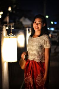 Portrait of smiling young woman standing against illuminated lamp