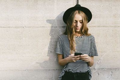 Young woman using phone while standing against wall