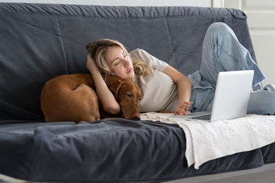 Woman with dog using laptop on sofa at home