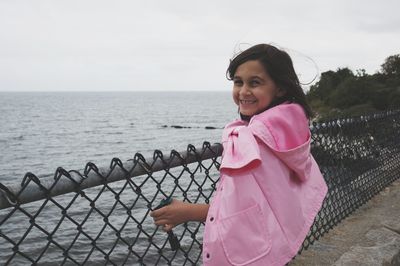 Smiling girl wearing pink coat standing by chainlink fence and sea against sky