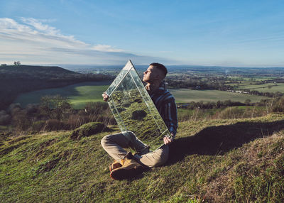 Young man holding mirror while sitting on landscape