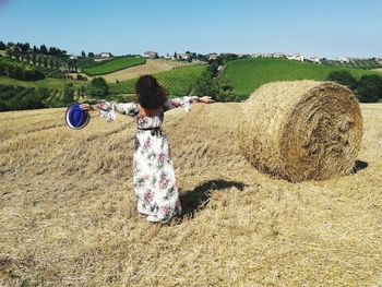 Rear view of woman with arms outstretched standing by hay bale on field