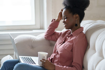 Side view of young woman holding coffee cup while sitting on sofa at home