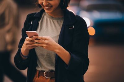 Midsection of smiling young woman using social media on phone while standing in city