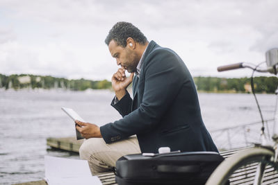 Side view of businessman looking at digital tablet while sitting on bench against bay