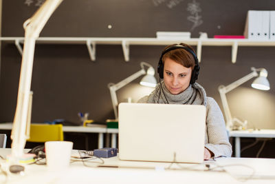 Confident businesswoman listening to headphones while using laptop at creative office