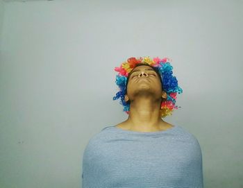 Young man wearing colorful wig while meditating against gray background